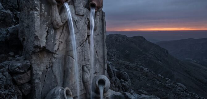 Gula, the Great One: a colossal weathered stone figure of a bearded Mesopotamian water-bearer standing astride a dark mountain ridge at dusk, two great vases raised overhead pouring bright streams of rain down toward the earth, two more vases at his