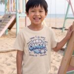 Cancer Aries: Street Sign × Scorpio: Street Sign Tail Badge Youth Classic Tee (Gildan 5000B), Natural, White in use: A beachside children's playground with a sandpit and pastel play equipment, soft sea-breeze daylight.