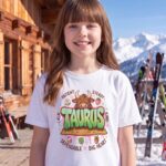 Taurus Street Sign Youth Classic Tee (Gildan 5000B), White, Sport Grey, Natural in use: Outside a cozy ski lodge on a bluebird day, snow-covered mountains and ski racks behind, crisp sunlight